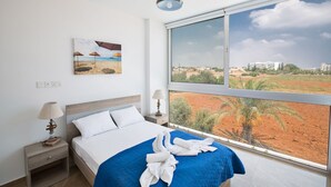 A bedroom with a bed, a nightstand, a lamp, a painting of a beach scene, and a large window with a view of a landscape.