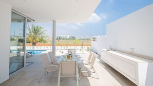 A modern outdoor patio with a dining table and chairs, a built-in kitchen counter, and a glass door leading to a pool area.