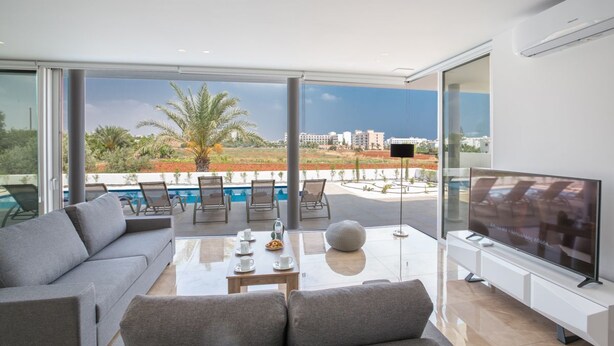 A modern living room with a large TV, a sofa, a coffee table, and a view of a pool and palm trees.
