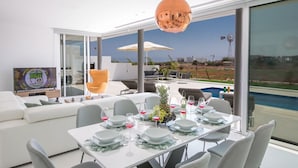 Een moderne eethoek met een tafel gedekt voor zes personen, een groot raam met uitzicht op een zwembad en een windmolen, en een televisie op de achtergrond.