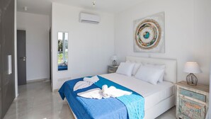 A modern hotel room with a large bed, a wall-mounted air conditioner, a decorative artwork, and a view of greenery through a window. - Villa Aglaia - Three Bedroom Villa, Sleeps 8 (Πρωταράς)