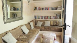 A living room with a brown sectional sofa, a small ottoman, a bookshelf with books, a decorative wall lamp, and a mirror.