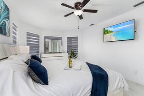 A modern bedroom with a bed, a ceiling fan, a TV, and a window with blinds. - Welcome to our Modern Cape Escape with Heated Pool (Cape Coral)