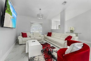 A modern living room with a white sofa set, a red accent chair, a white coffee table, and a large flat-screen TV mounted on the wall.