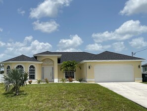 A two-story house with a garage, a front yard with grass and small plants, and a clear blue sky with clouds. - Welcome to our Modern Cape Escape with Heated Pool (Cape Coral)