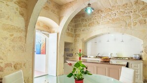 A dining area with a glass table, white chairs, a red potted plant, and a kitchen in the background with white cabinets and a stone wall.