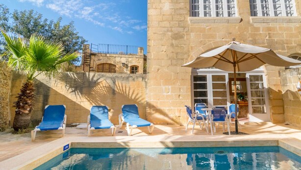 A pool area with lounge chairs and an umbrella, a stone building with arched windows, and a palm tree.
