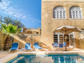 A pool area with lounge chairs and an umbrella, a stone building with arched windows, and a palm tree. - Farmhouse Bertu - Two Bedroom Villa, Sleeps 4 (Xagħra)