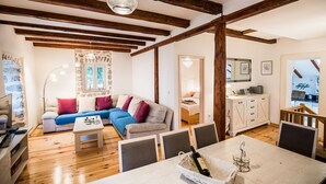 Un accogliente soggiorno con soffitto in legno, un divano con cuscini, un tavolo da pranzo con sedie e una zona cucina visibile attraverso un'apertura.