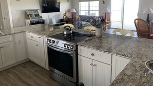 A modern kitchen with granite countertops, white cabinetry, a built-in oven, and a dining area with a striped sofa and a large window. - Gulf to Bay, on the beach home with bay access (Alligator Point)