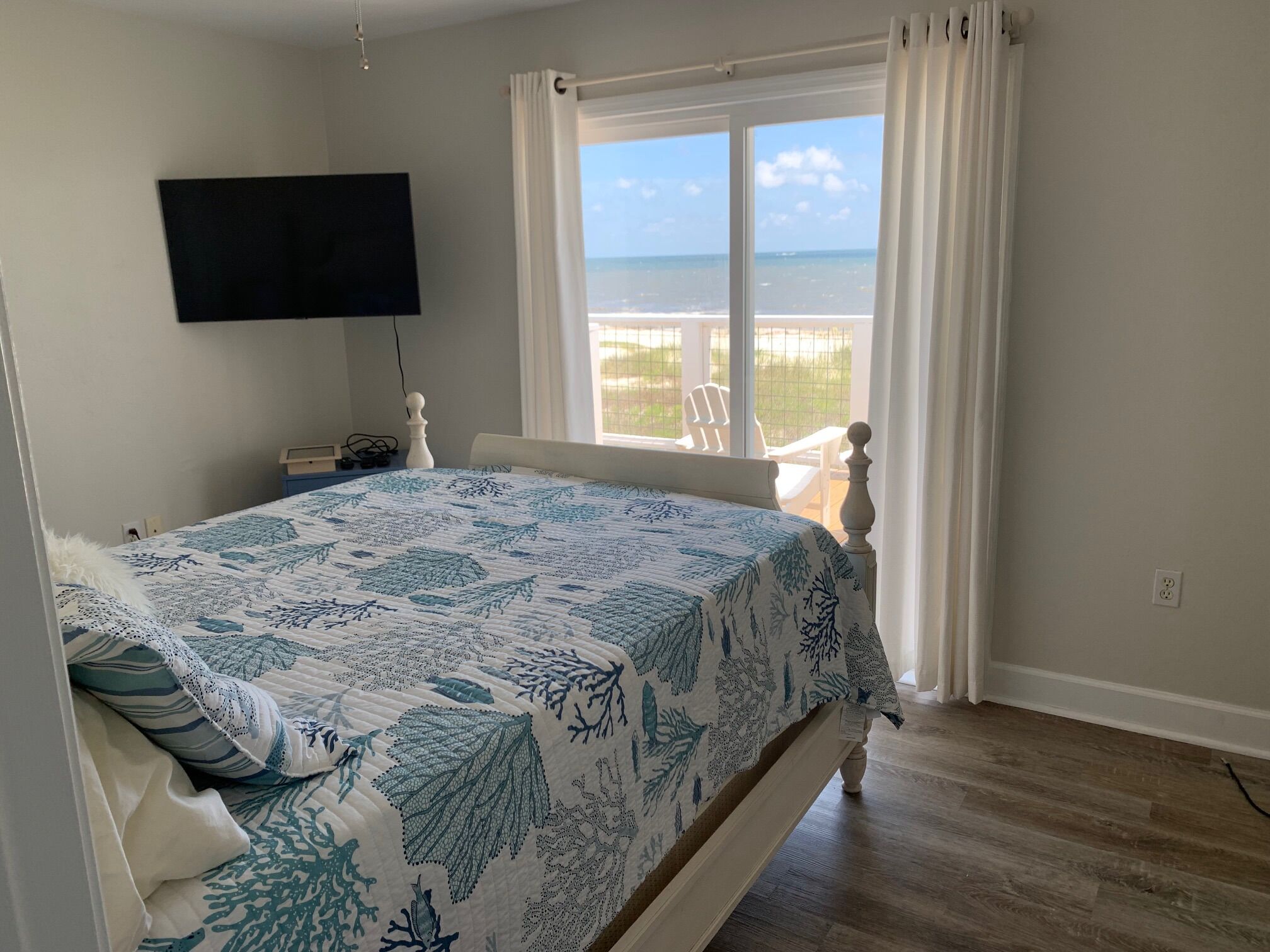 Een slaapkamer met een bed, een aan de muur bevestigde televisie, een raam met gordijnen en uitzicht op de oceaan.