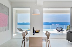 Un comedor moderno con una mesa, sillas y una lámpara colgante. Hay un balcón con vistas al mar.