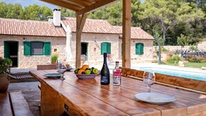 A wooden table set for dining outdoors with a view of a stone house with green shutters, a swimming pool, and trees in the background. - Old Town Villa Hvar - Five Bedroom Villa, Sleeps 12 (Uvala Tatinja)