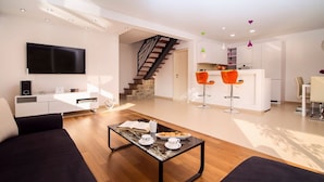 A modern living room with a sofa, a coffee table with pastries, a TV, and a staircase leading to a kitchen area.