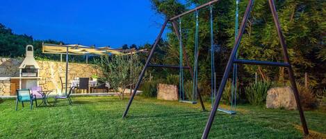 A backyard with a swing set, a barbecue area, and a stone wall.