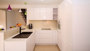 A modern kitchen with white cabinetry, a black sink, a built-in oven, and a microwave. There are colorful pendant lights and a wooden ladder against the wall.
