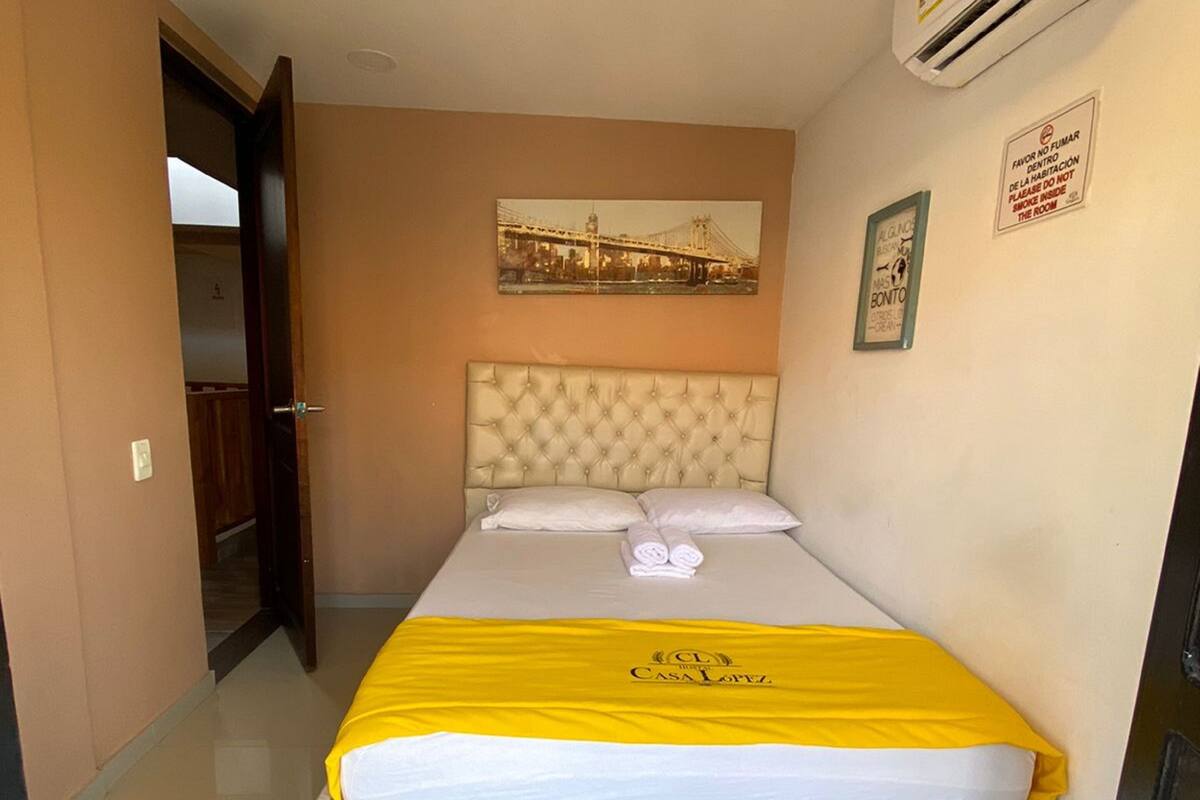 A hotel room with a bed, a headboard, a wall-mounted air conditioner, a framed picture, and a sign.