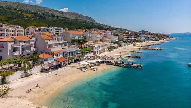 A coastal town with buildings along the beach, clear blue waters, and a mountainous backdrop.