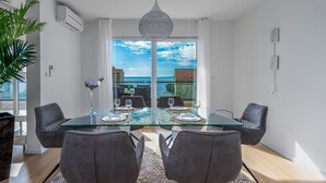 A modern dining area with a glass table, grey upholstered chairs, and a view of the sea through large windows.