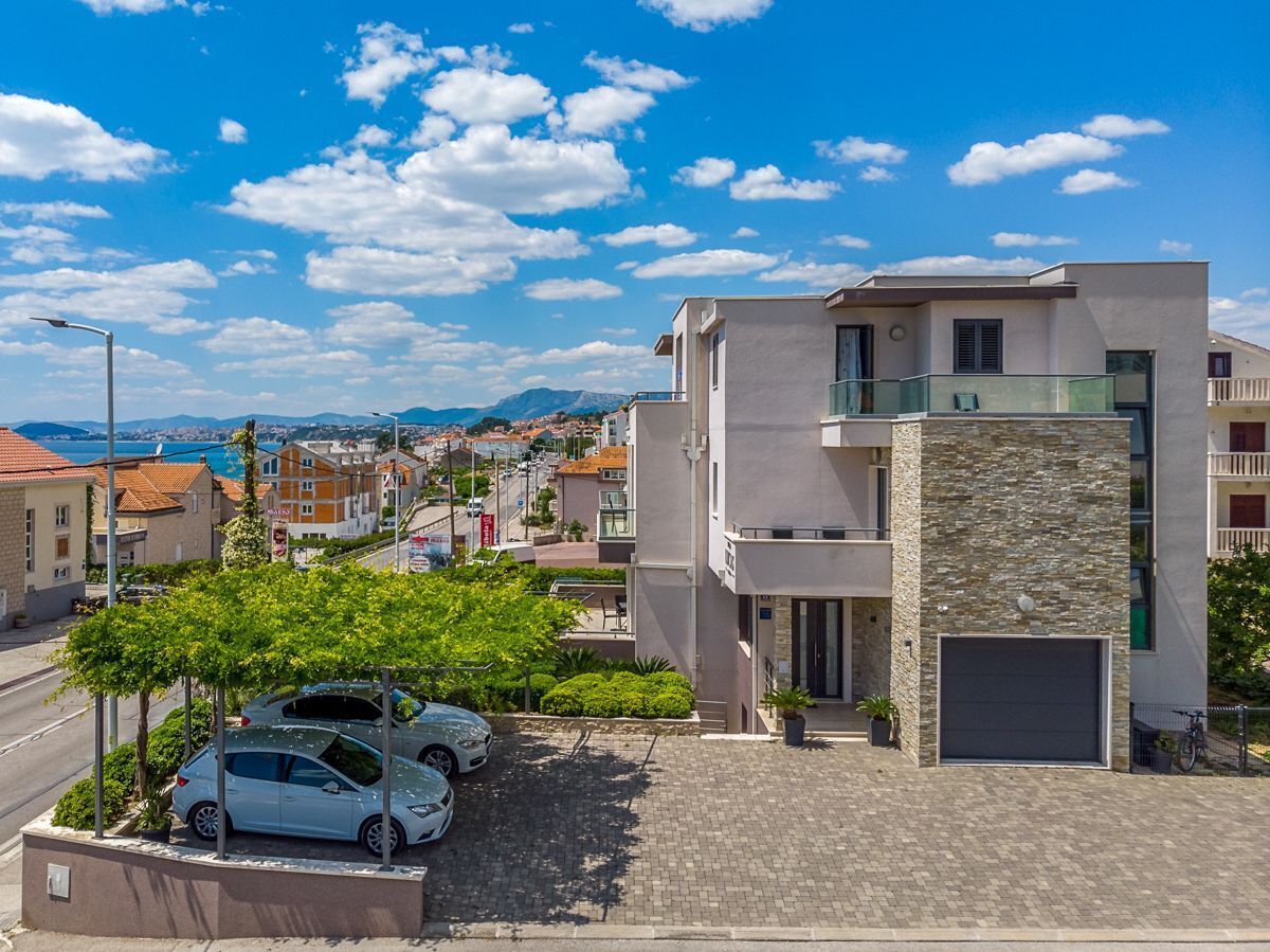 A modern two-story building with a stone facade, a covered parking area, and a view of the sea and mountains in the background.
