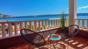 A balcony with two black wire chairs, a small round table, and a drink. The balcony overlooks a body of water and a distant coastline.
