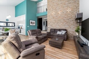 A modern living room with a sectional sofa, a coffee table, and a TV stand. There is a kitchen area in the background with a dining table and chairs.