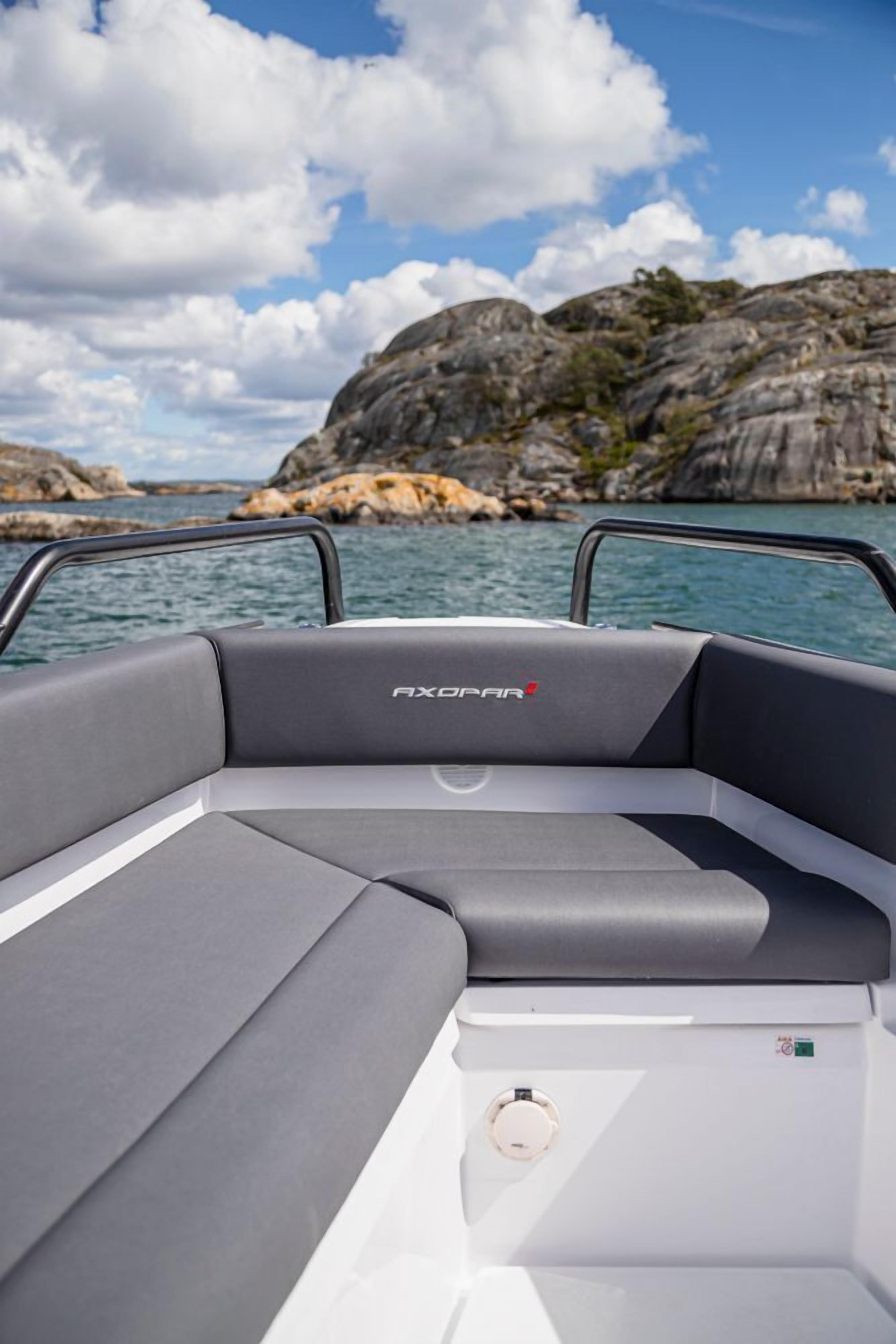 A boat interior with a gray cushioned seating area, a white control panel, and a view of rocky coastline and water.