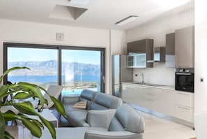 Een moderne woonkamer met een grijze bank en een eettafel, een keuken met witte kasten, en een groot raam met uitzicht op bergen.