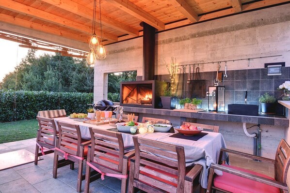 An outdoor dining area with a wooden table and chairs, a built-in grill, and a stone wall backdrop. - Villa Drvo - Five Bedroom Villa, Sleeps 10 (Smoljanci)