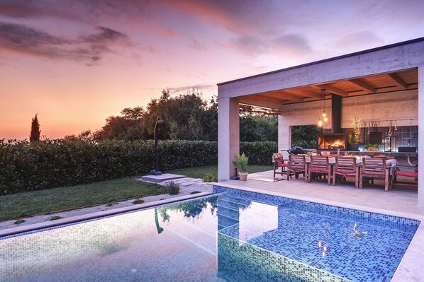 A modern outdoor pool area with a dining setup and a view of the sunset.