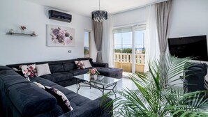 A modern living room with a black sofa, a glass coffee table, and a large window with a view of buildings. - Villa Novi - Five Bedroom Villa, Sleeps 12 (Rudine)