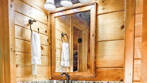 Un baño estilo cabaña de madera con encimera de granito, un lavabo con grifo y un espejo encima. Hay armarios de madera debajo del espejo y una toalla colgada de un gancho.