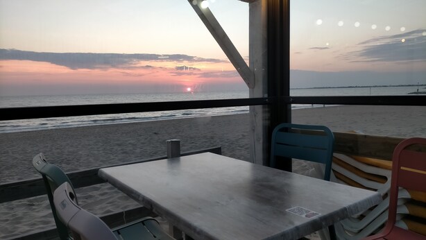 Un restaurante frente a la playa con una mesa y sillas, con vista al océano y al atardecer.