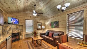 A cozy cabin interior with a stone fireplace, wooden walls, a leather sofa, a dining table, and a ceiling fan. - Hook, Line & Sinker: Perfect Cabin Nestled in Woods Only Minutes from Gatlinburg Strip (Gatlinburg)