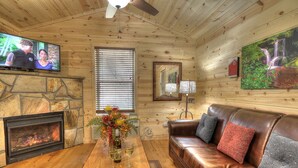 A cozy cabin interior with a stone fireplace, a leather sofa, a wooden coffee table with a vase of flowers, a television mounted on the wall, and a painting of a waterfall. - Hook, Line & Sinker: Perfect Cabin Nestled in Woods Only Minutes from Gatlinburg Strip (Gatlinburg)