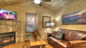 A cozy cabin interior with a stone fireplace, leather sofa, wooden flooring, a ceiling fan, a TV, and a painting of a landscape. - Fish Tales: Cozy Cabin Perfect for Family Getaway (Gatlinburg)