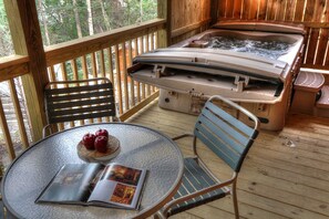 Una terraza de madera con un jacuzzi, dos sillas, una mesa con una revista y manzanas, y vistas a árboles.