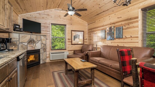 Een gezellig cabin interieur met een stenen open haard, houten wanden, een bruine leren bank, een houten salontafel en een plafondventilator.
