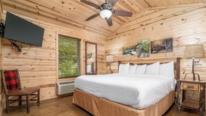 Een houten slaapkamer met een bed, een stoel, een plafondventilator, een televisie en een raam met jaloezieën.