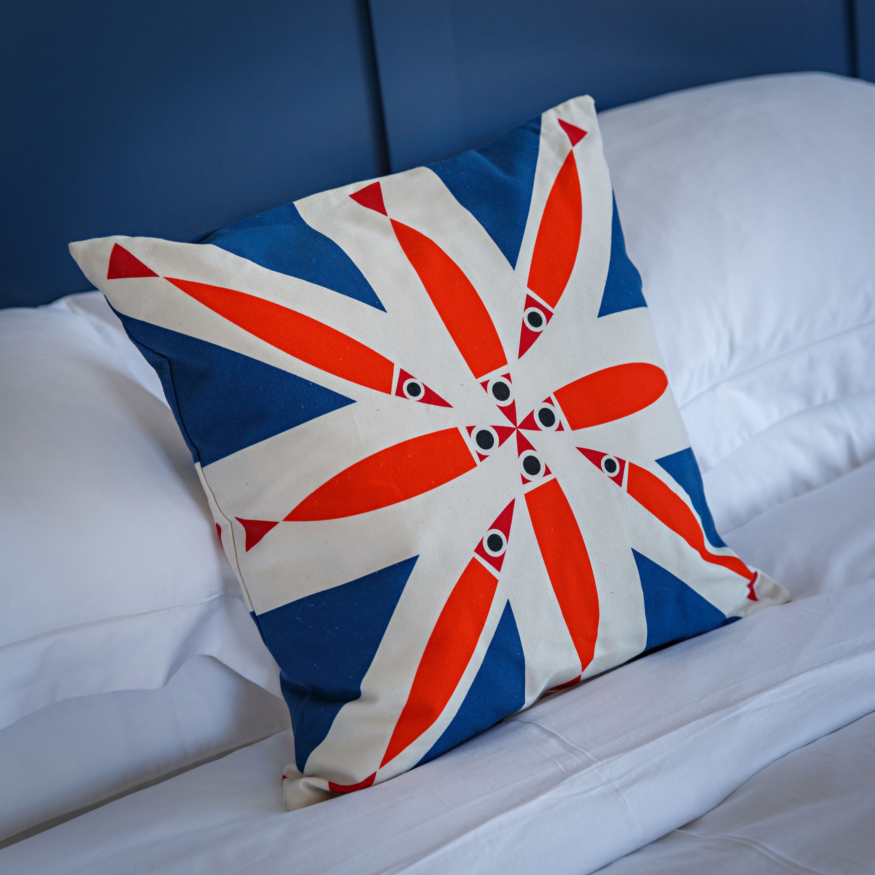 Almohada con diseño de la bandera del Reino Unido sobre una cama con sábanas blancas y cabecera azul.