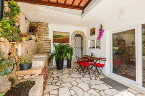 Un patio de piedra con una mesa y sillas, un muro de piedra con plantas y vistas a la cocina a través de un vidrio.