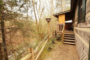 Una cabana de fusta amb una escala de fusta que condueix a una terrassa, envoltada d'arbres i un rierol.