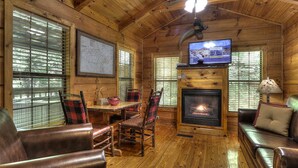 Ein gemütliches Cabin-Interieur mit Holzbalkendecke, Kamin, einem darüber montierten Fernseher und einem Essbereich mit Stühlen und Tisch.