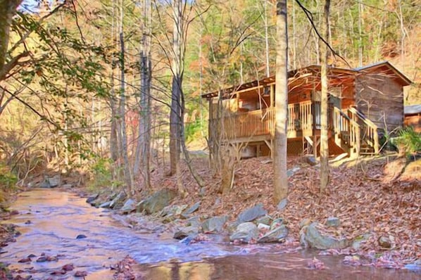 Uma cabana com deck de madeira e escadas para um alpendre, rodeada por árvores e um riacho.