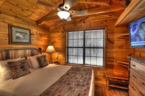 Quarto de cabana de madeira com cama, ventoinha no teto, janela com persianas, quadro pendurado na parede e televisão mostrando a previsão do tempo.