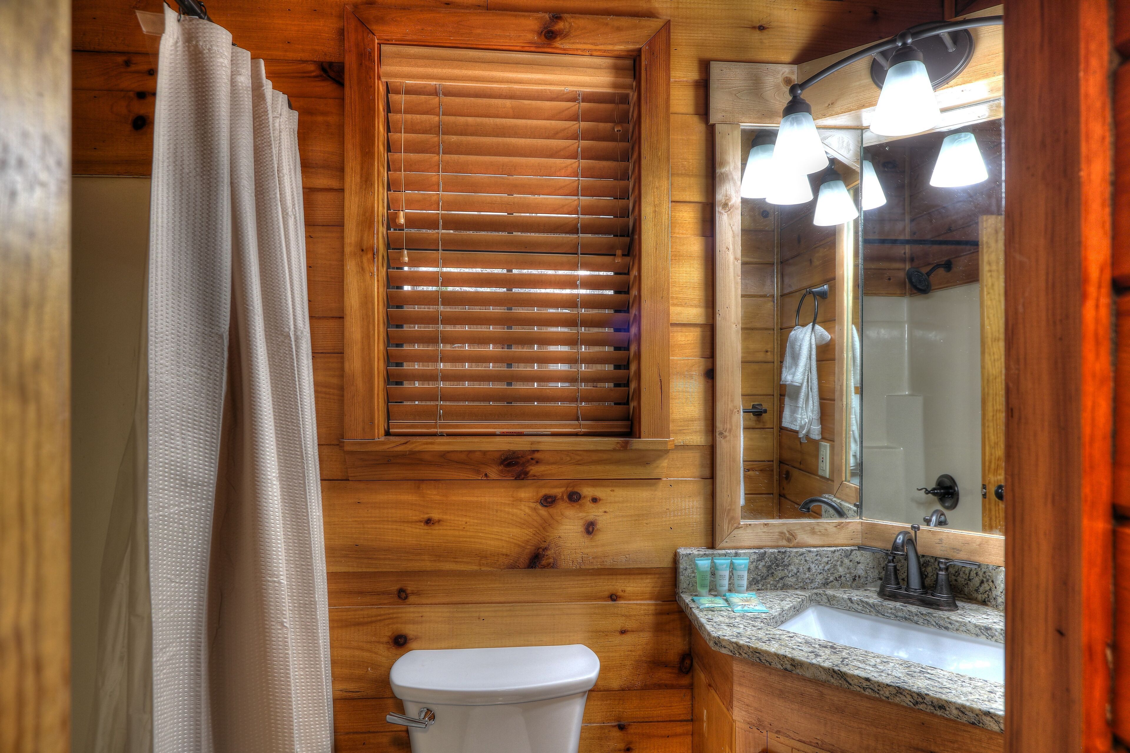 A wooden bathroom with a toilet, sink, and window with blinds.