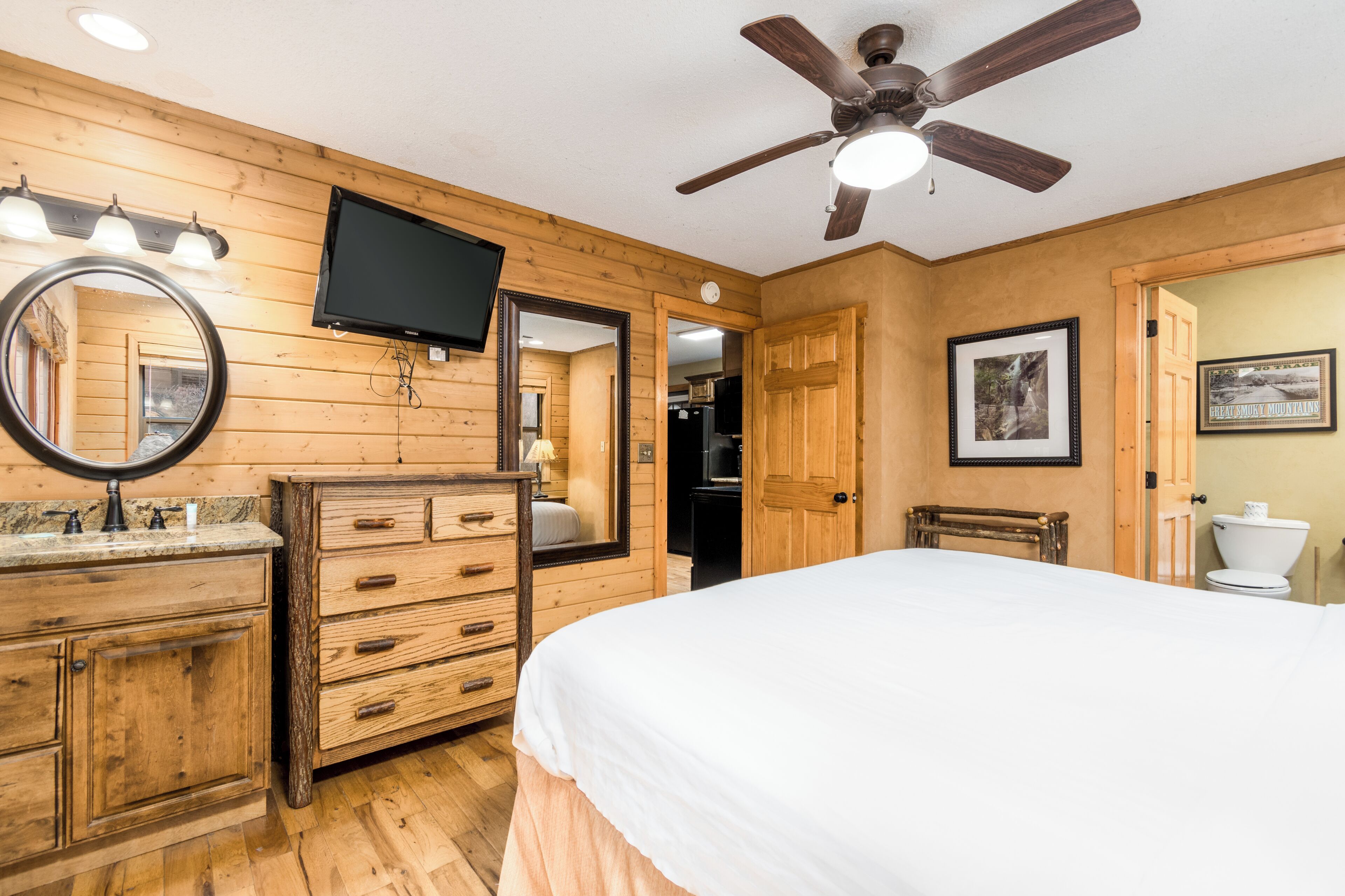 A wooden-paneled room with a bed, dresser, mirror, ceiling fan, and a TV.