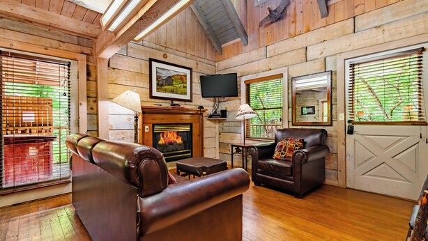 Interior acogedor de una cabaña de troncos con chimenea, sofás de cuero, pisos de madera y ventanas amplias con persianas.