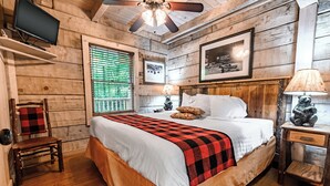 Un dormitorio tipo cabaña de madera con cama, televisor en un estante, una silla y una ventana con persianas.