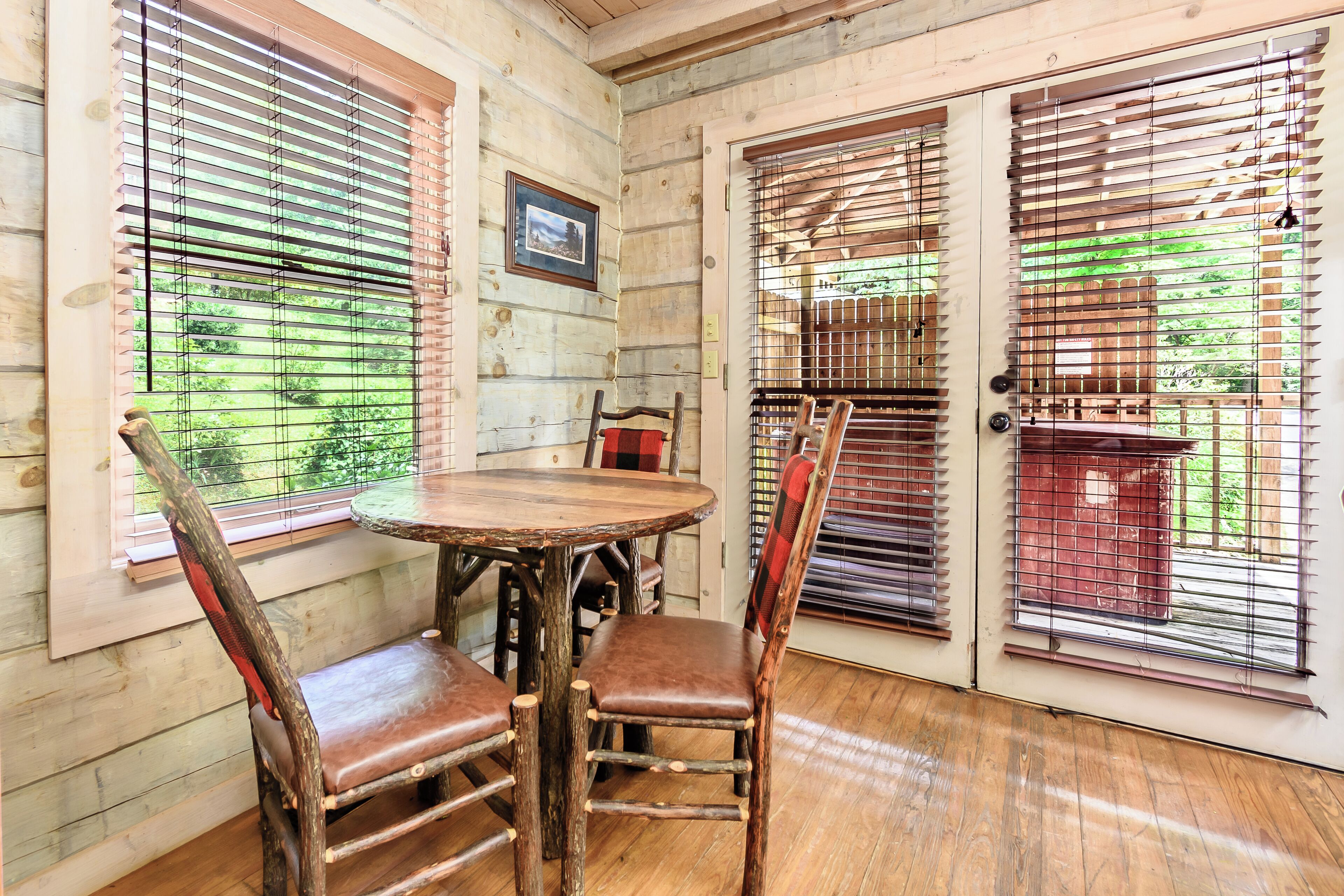 Un intérieur en bois avec une table ronde et quatre chaises, de grandes fenêtres équipées de stores et une porte avec un portail en métal.